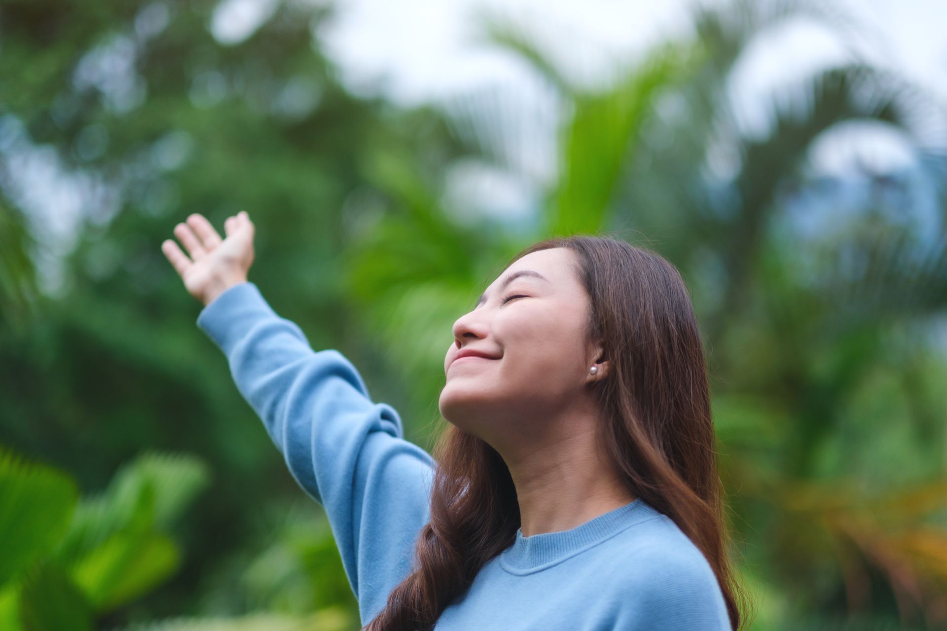 A person wearing a blue sweater stands outdoors in nature with their eyes closed, smiling, and one arm raised upward.