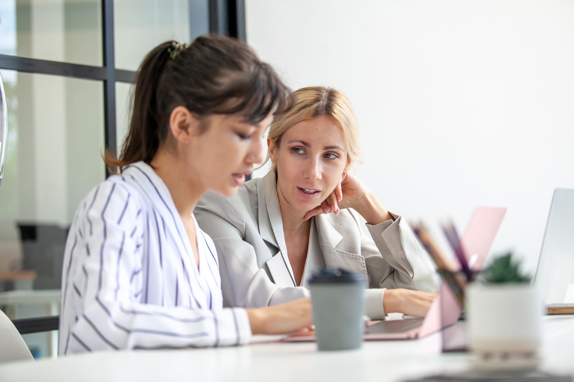 Two colleagues sitting at a white desk in a bright office, one speaking and the other listening while looking at a laptop.