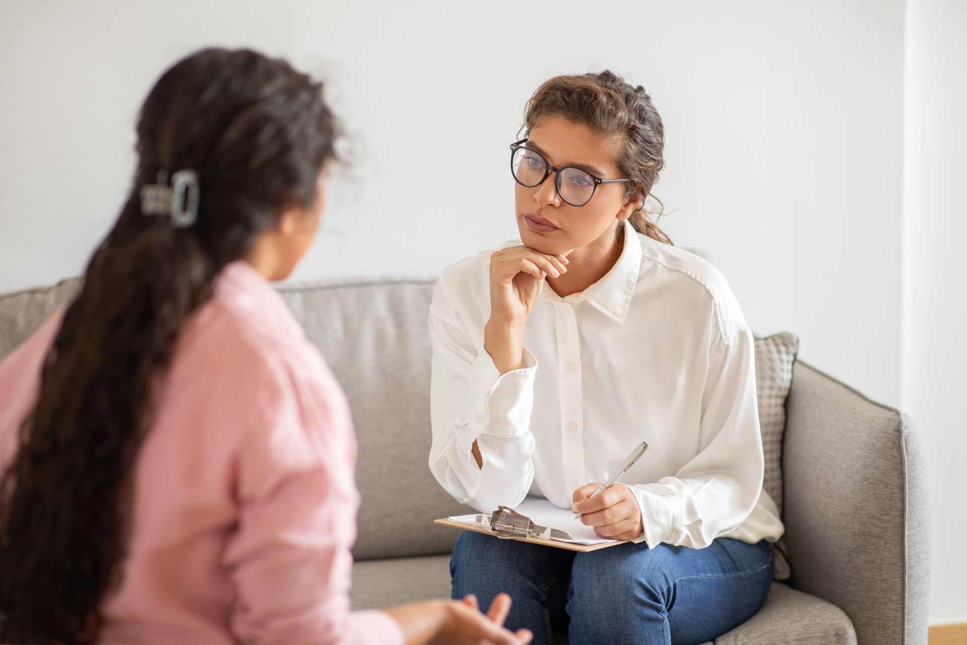 A person in a pink shirt sits on a sofa, speaking to a person in a white shirt who is listening with a notepad on a couch.