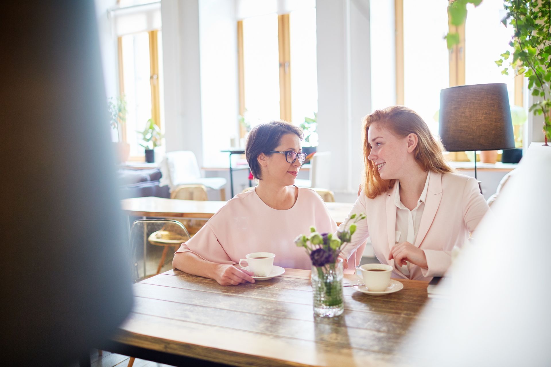 Two people sitting at a wooden cafe table, talking and holding coffee cups in a brightly lit room.