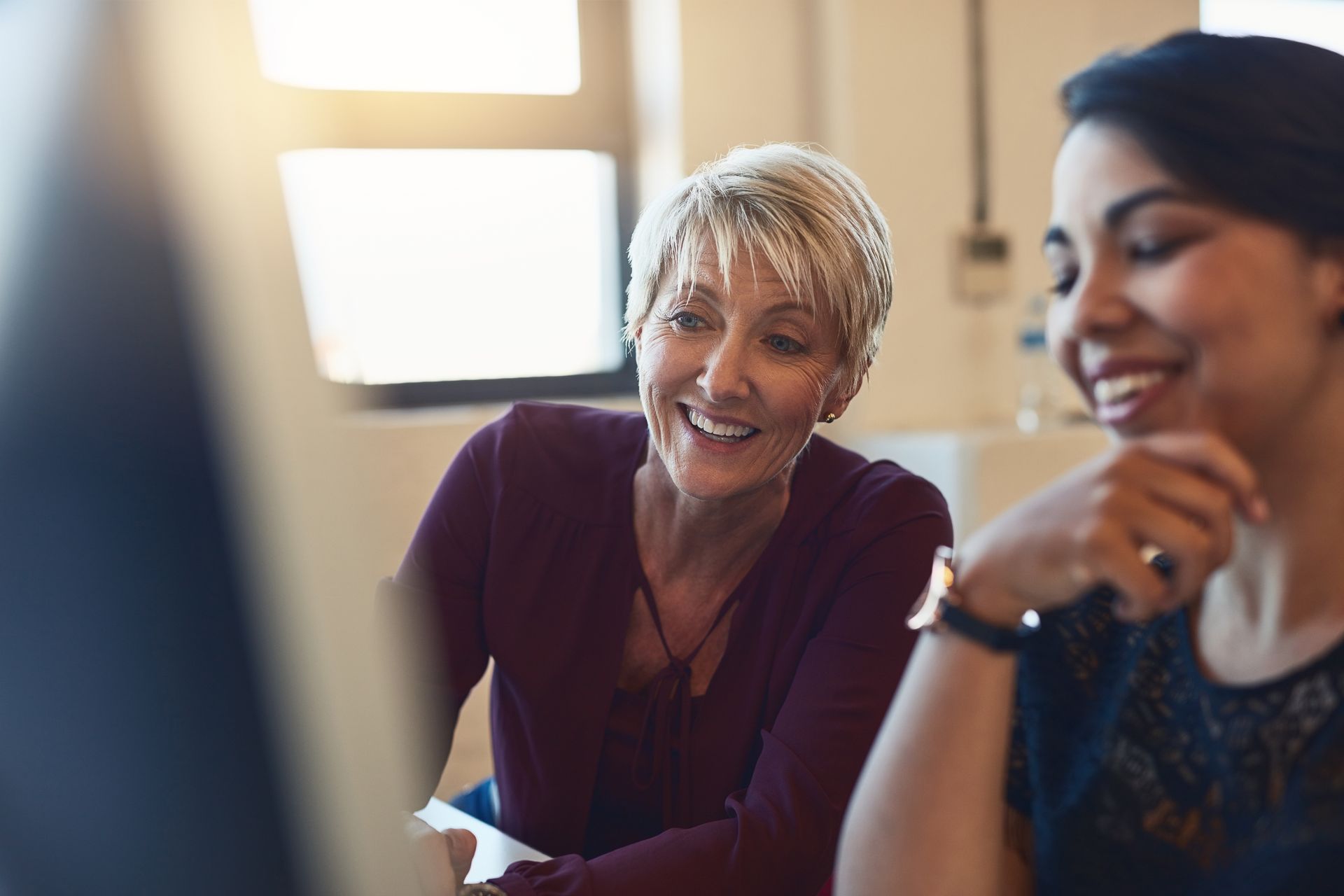 Two colleagues smiling while working together on a computer in a brightly lit office.