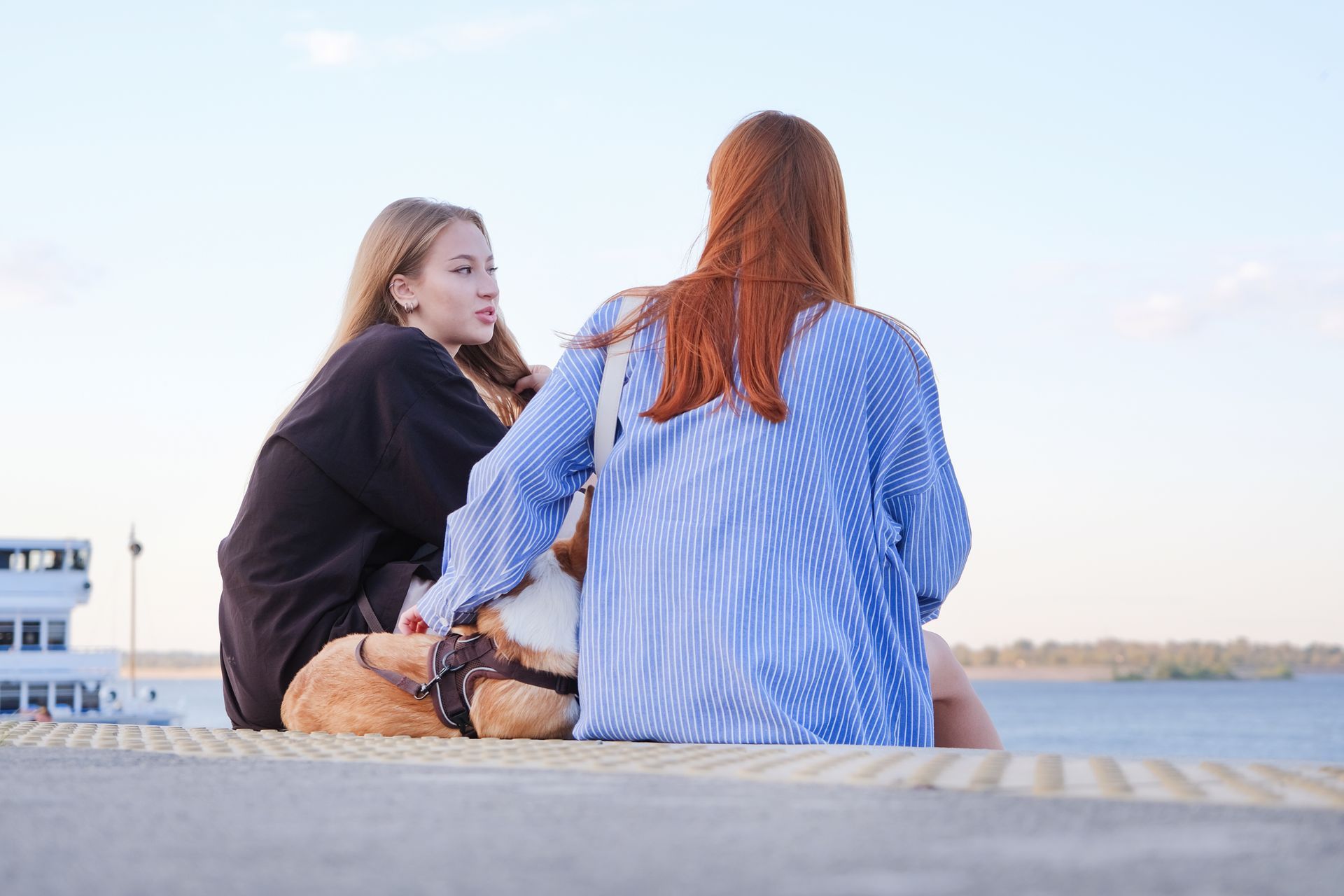 Two people sit on a concrete pier by the water with a dog, one person turned to look at the other.