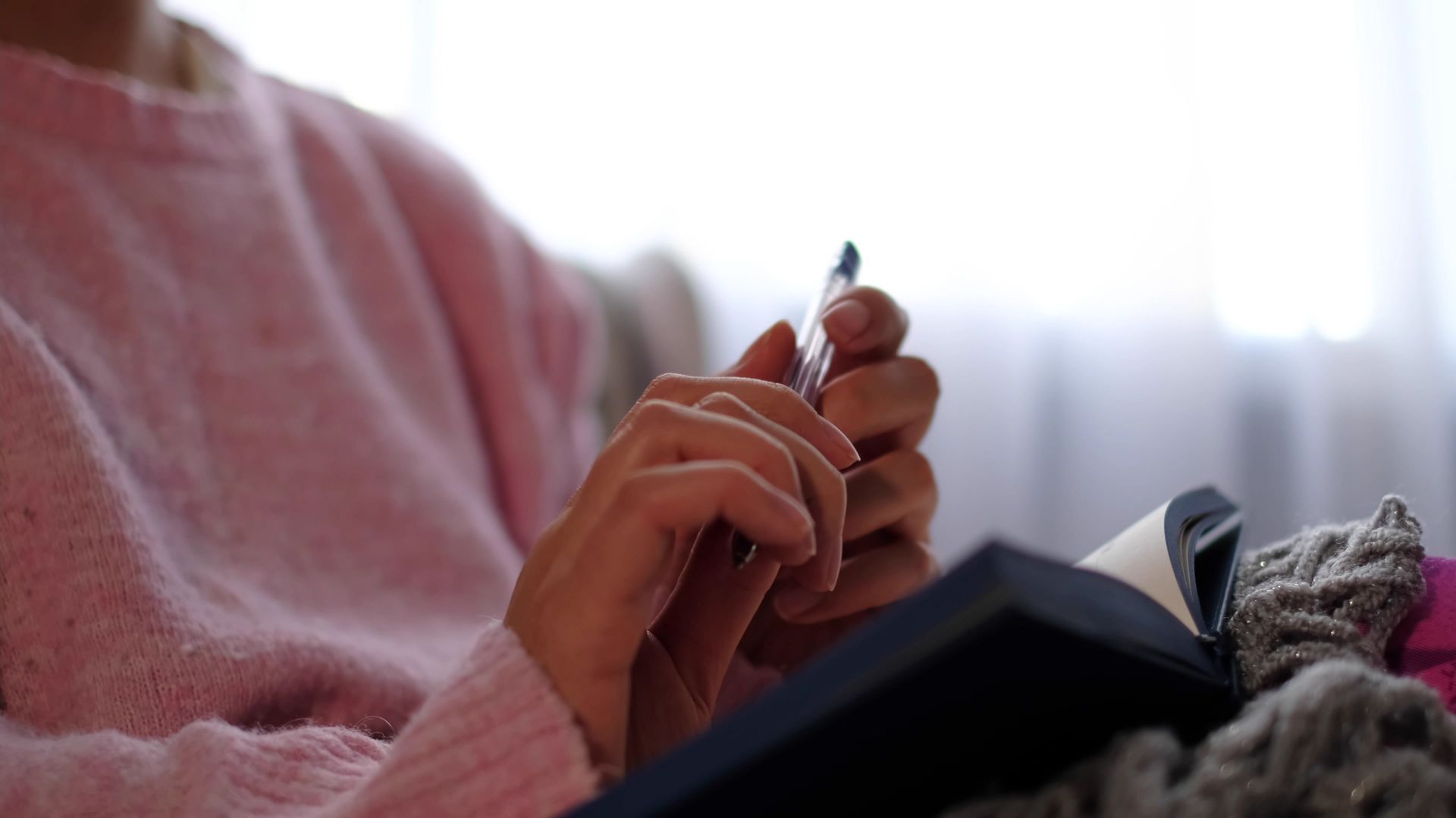 A person in a pink sweater holds a pen, poised to write in an open notebook while resting on a soft surface.