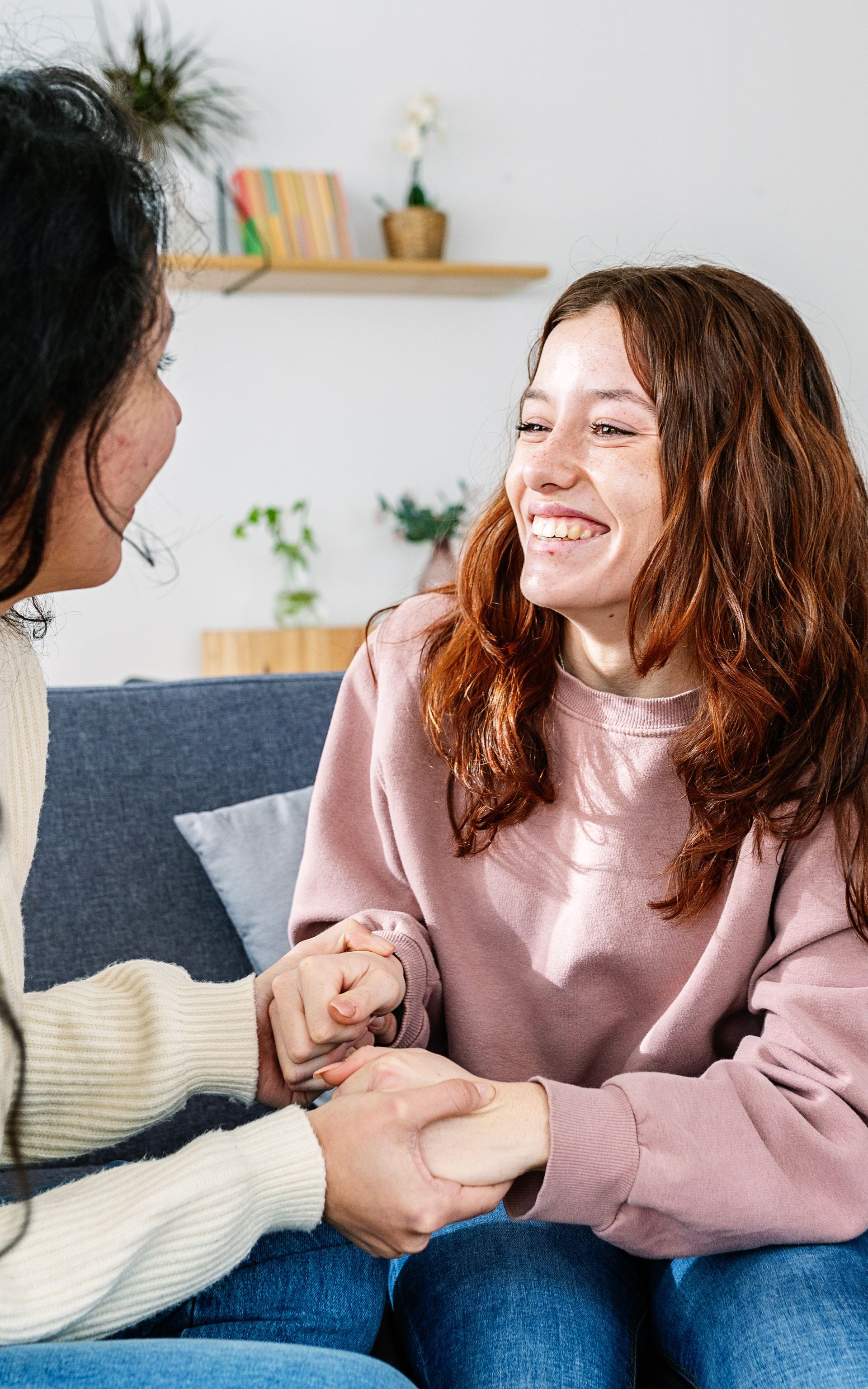 Two people holding hands and smiling while sitting on a sofa in a brightly lit room with a shelf in the background.