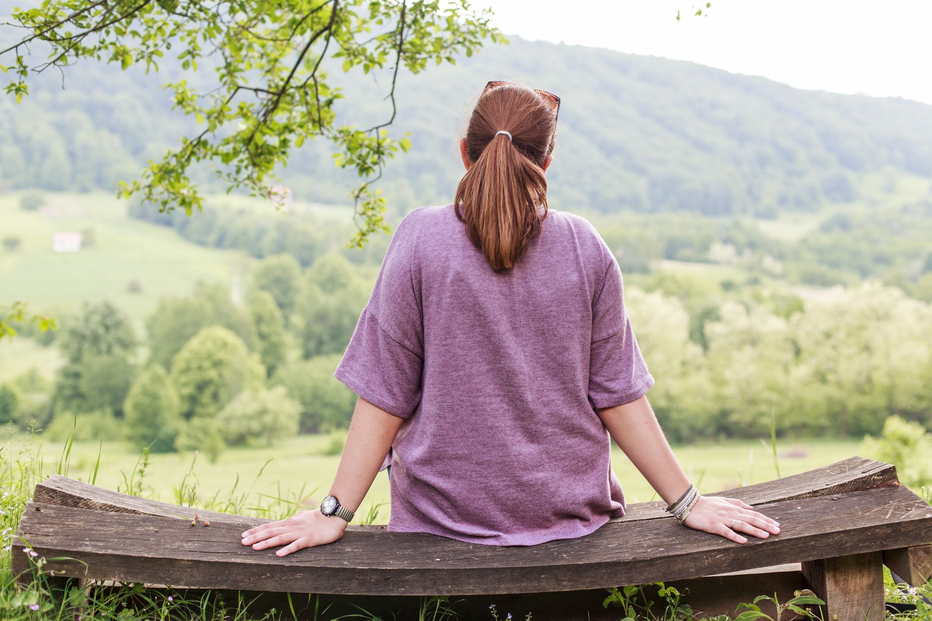 A person with a ponytail, wearing a purple shirt, sits on a rustic wooden bench overlooking a lush green hilly landscape.