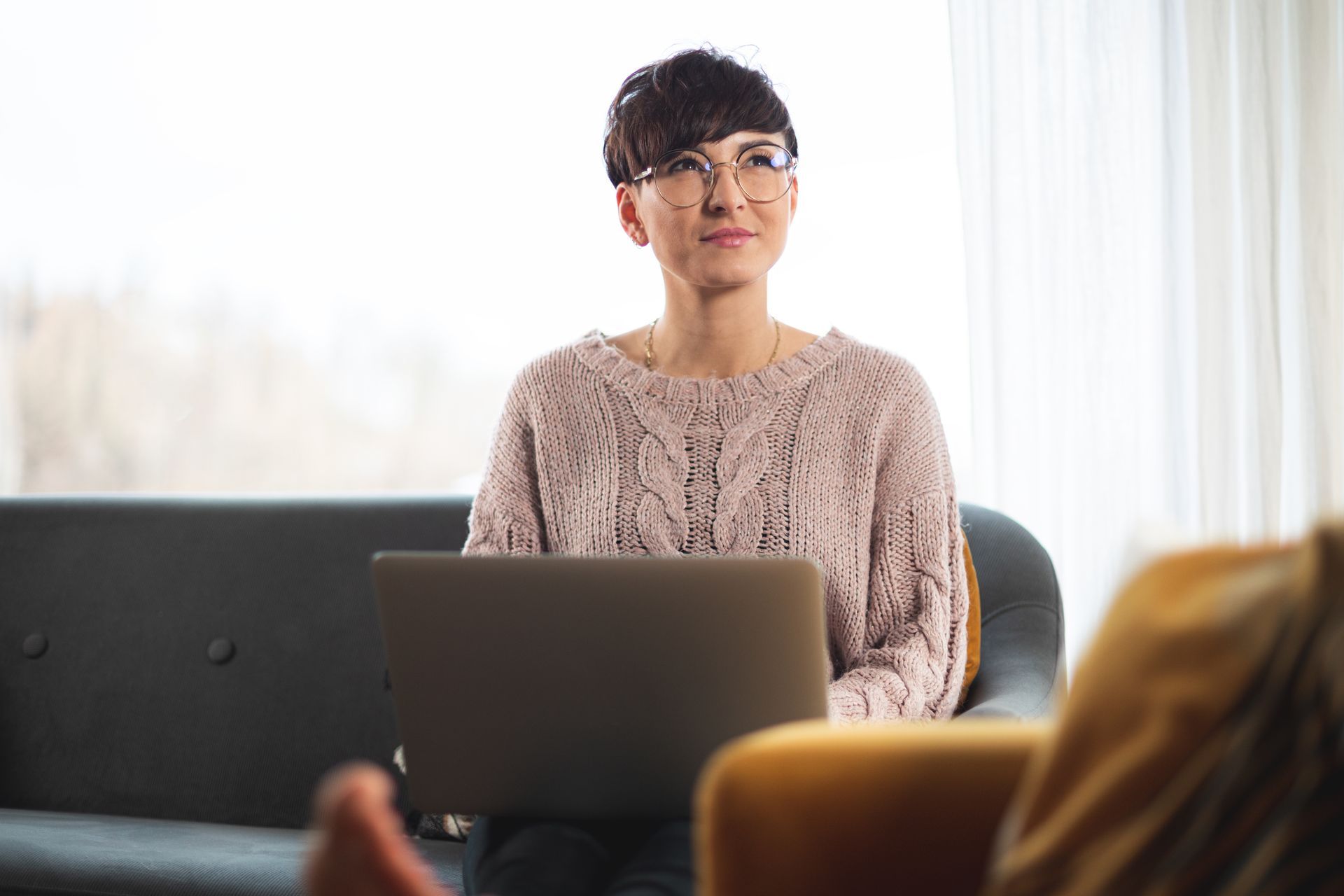 A person wearing glasses and a pink knit sweater sits on a dark sofa with a laptop, looking upward in a bright room.