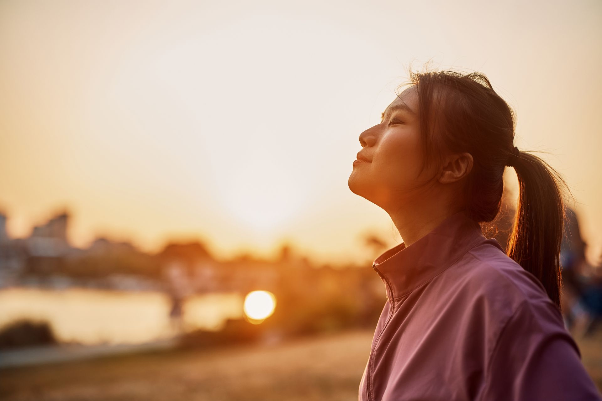A person with a ponytail stands outdoors at sunset, eyes closed and looking upward, wearing a mauve jacket.