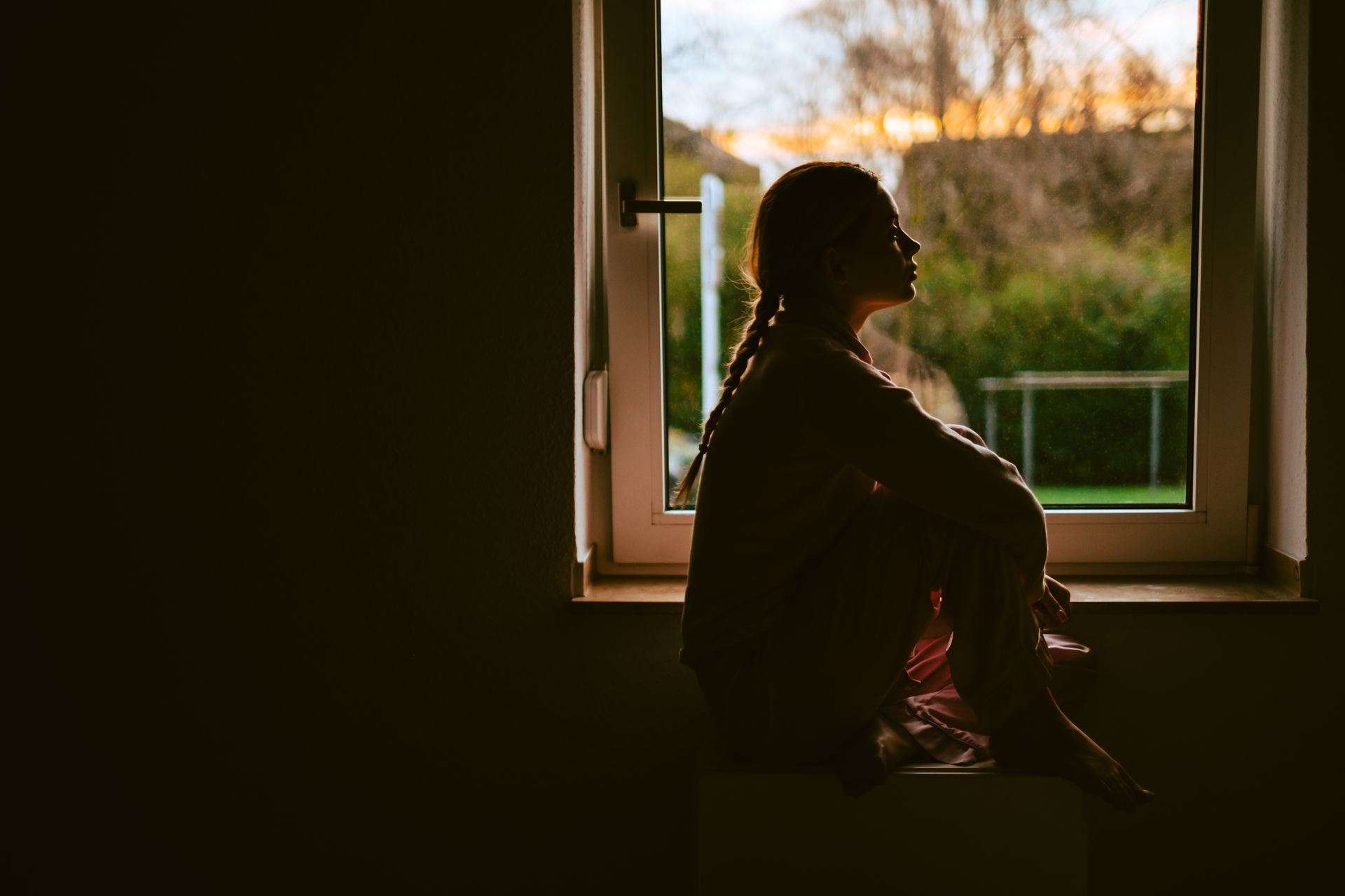 A person sits in silhouette on a window ledge, gazing out at the late afternoon light.