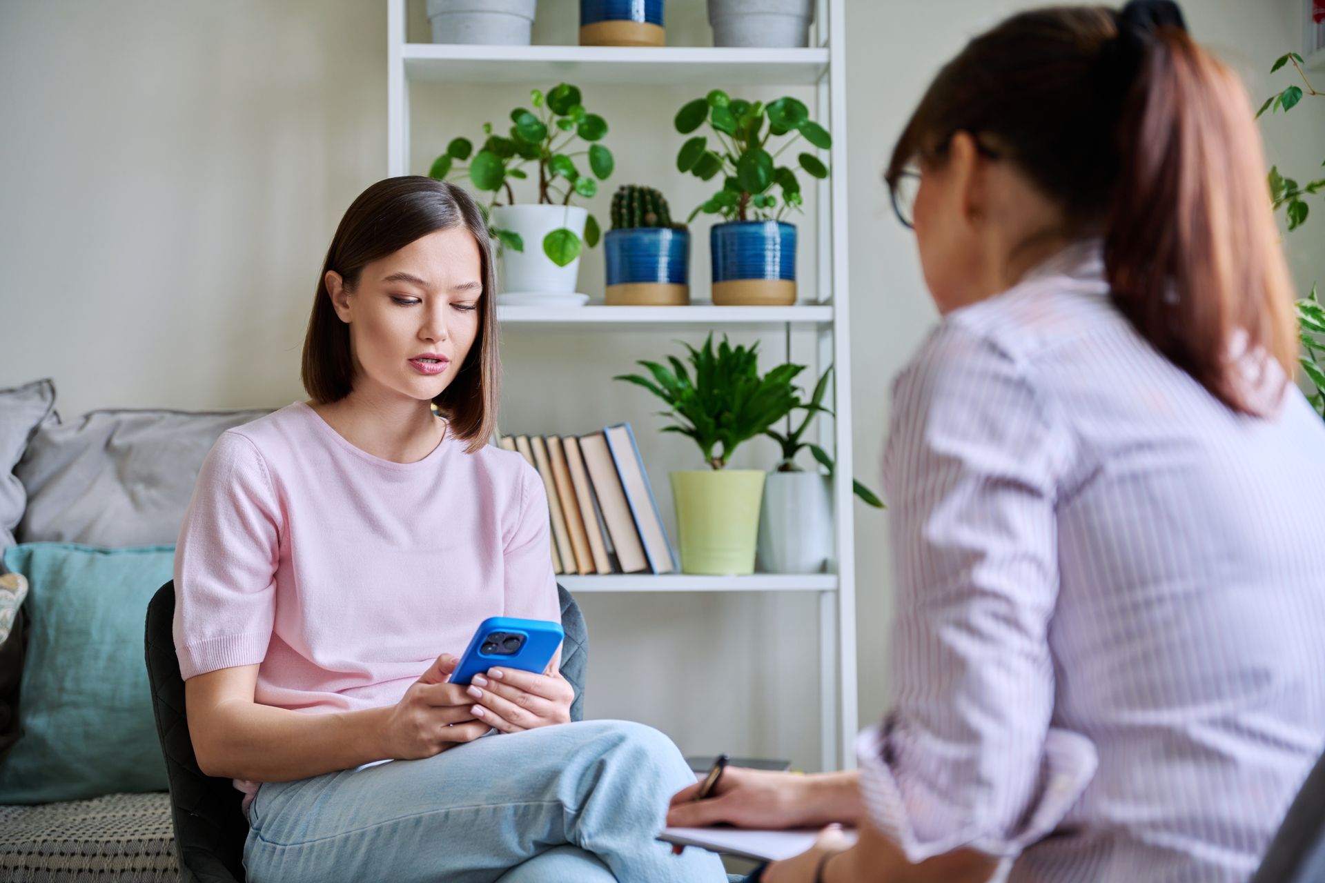 A person looks at their phone while sitting across from a professional taking notes in a brightly lit office with plants.