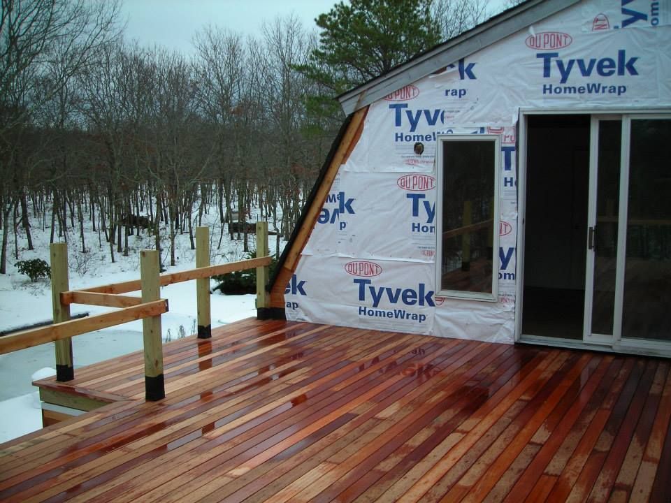 Tyvek-wrapped house under construction with a wet wooden deck and snowy woods beyond