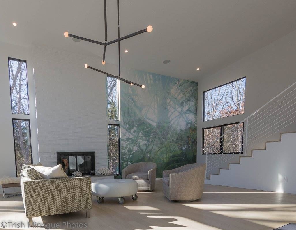 Modern living room with tall windows, white staircase, patterned chairs, and abstract wall art