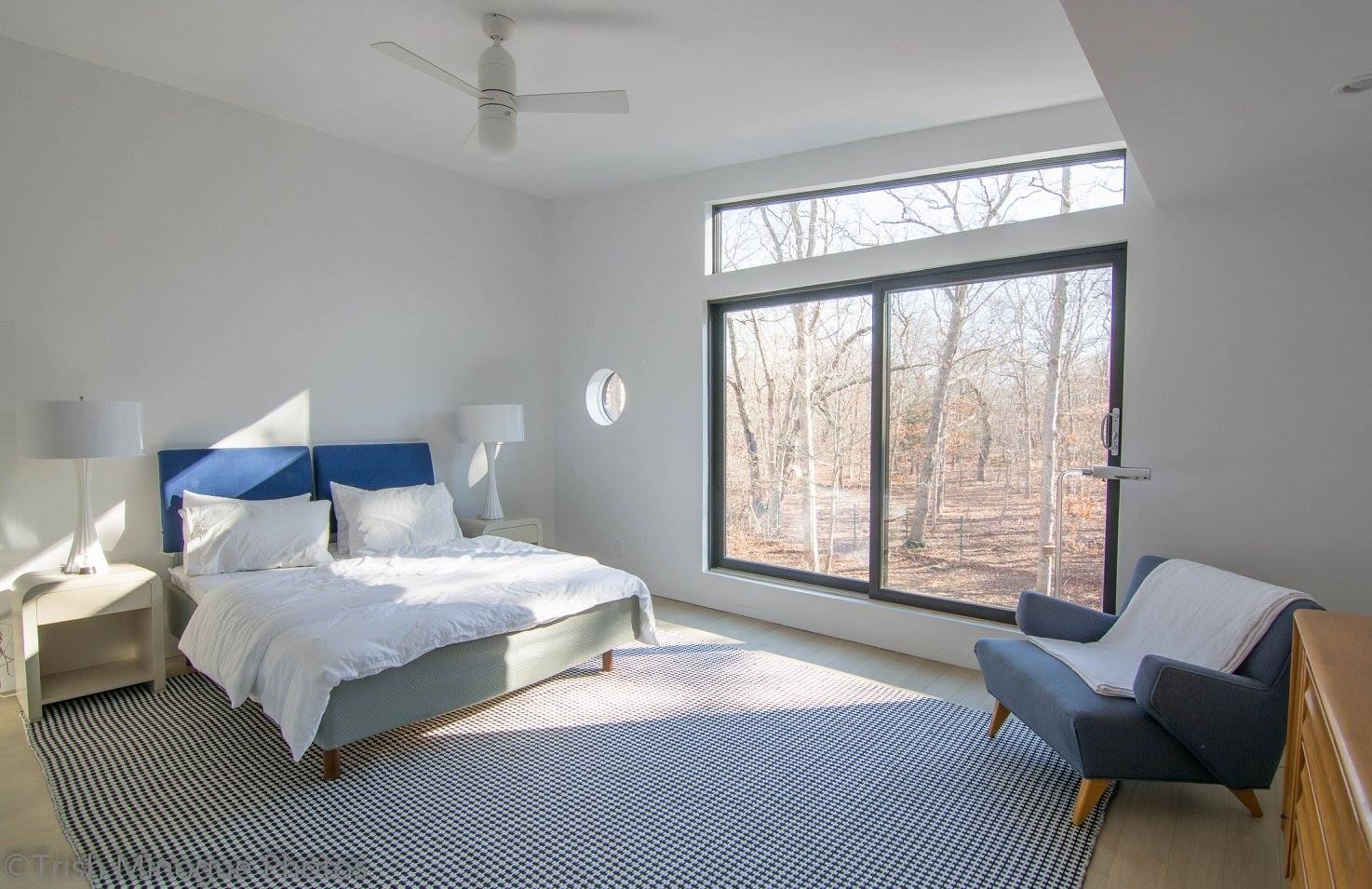 Bright bedroom with large window, blue accent chair, bed, and patterned rug in a minimalist white room