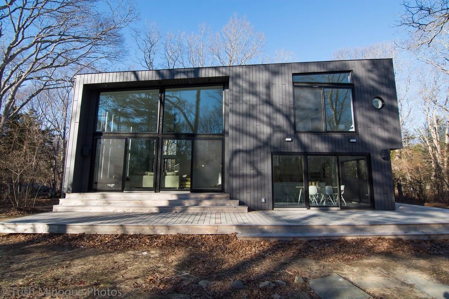Modern two-story dark gray house with large windows and a front patio in a wooded setting