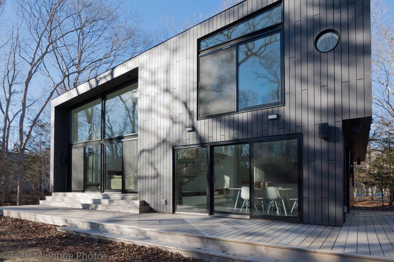 Modern two-story house with large glass windows and dark exterior panels in a wooded setting