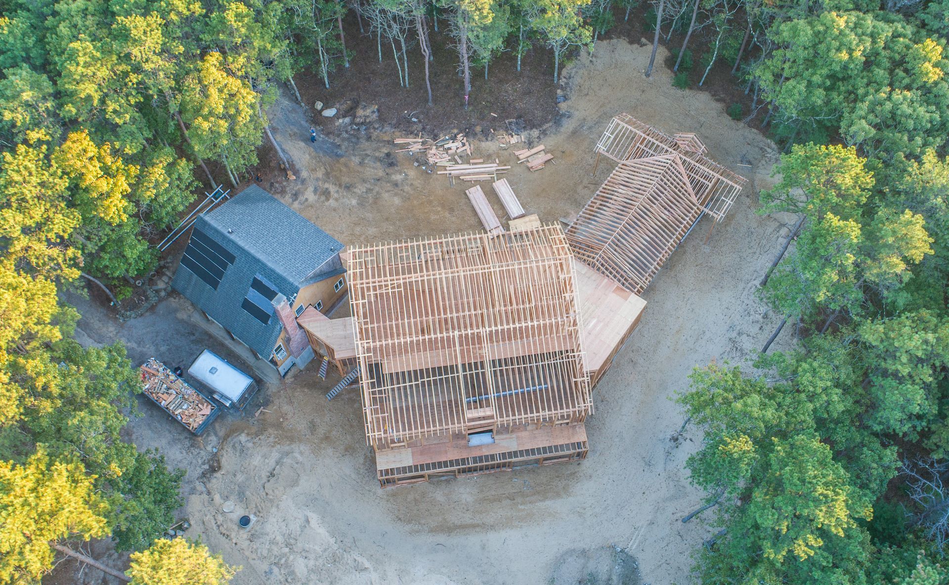 Aerial view of a house under construction beside a blue shed, surrounded by trees and dirt clearing