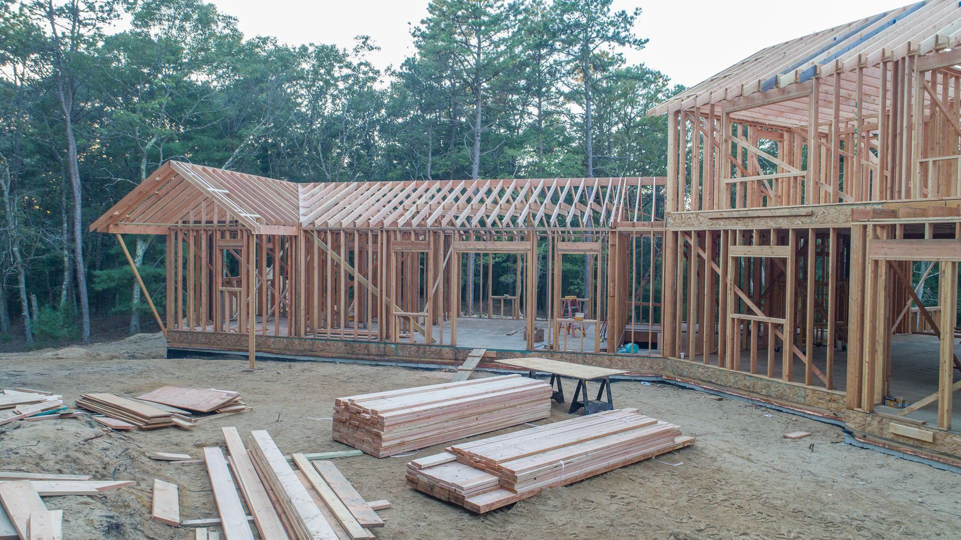 Wood-framed houses under construction with lumber stacks and a dirt foundation in a wooded area
