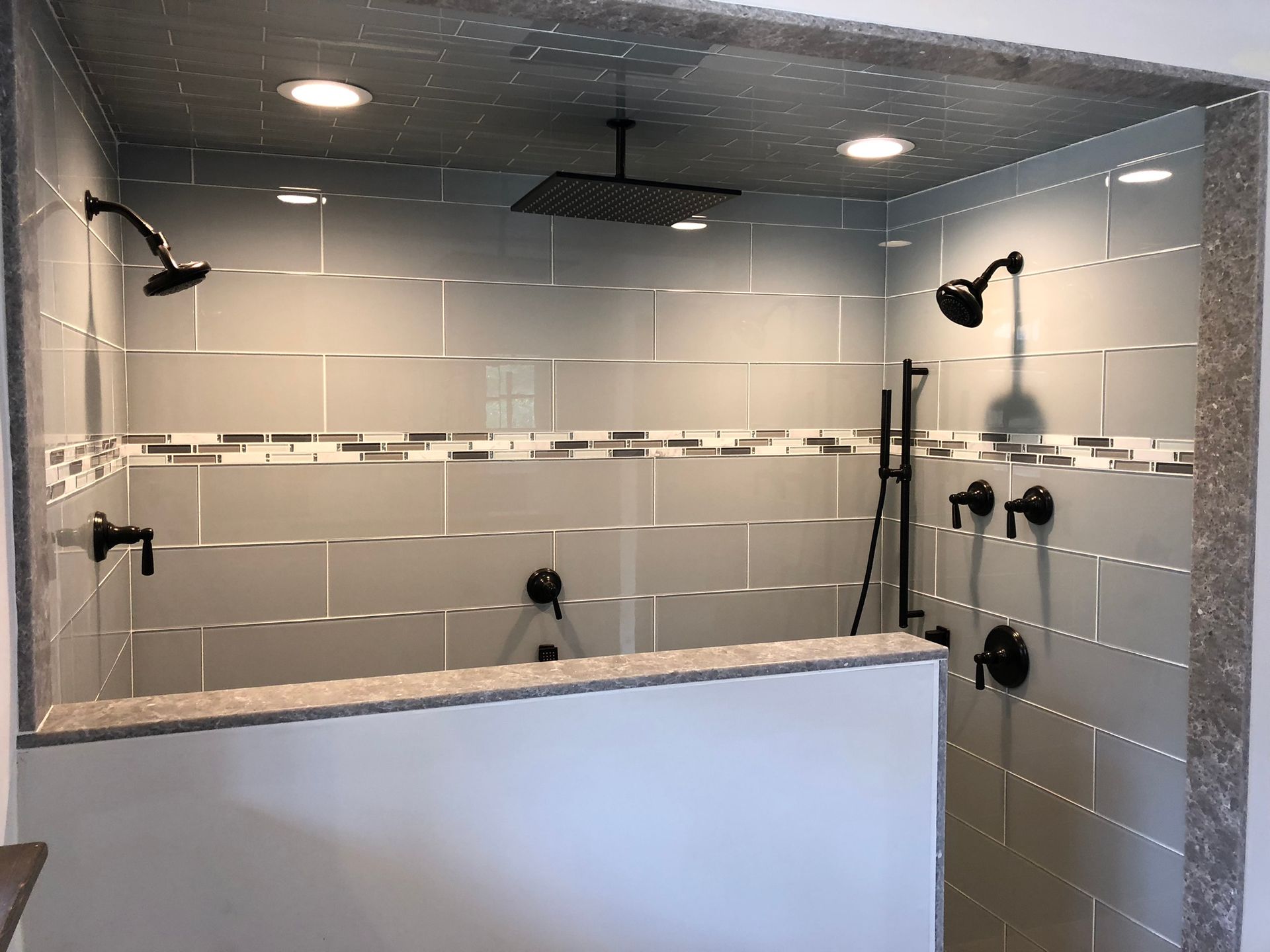 Modern tiled shower with multiple black showerheads and a half-wall, viewed from the bathroom side