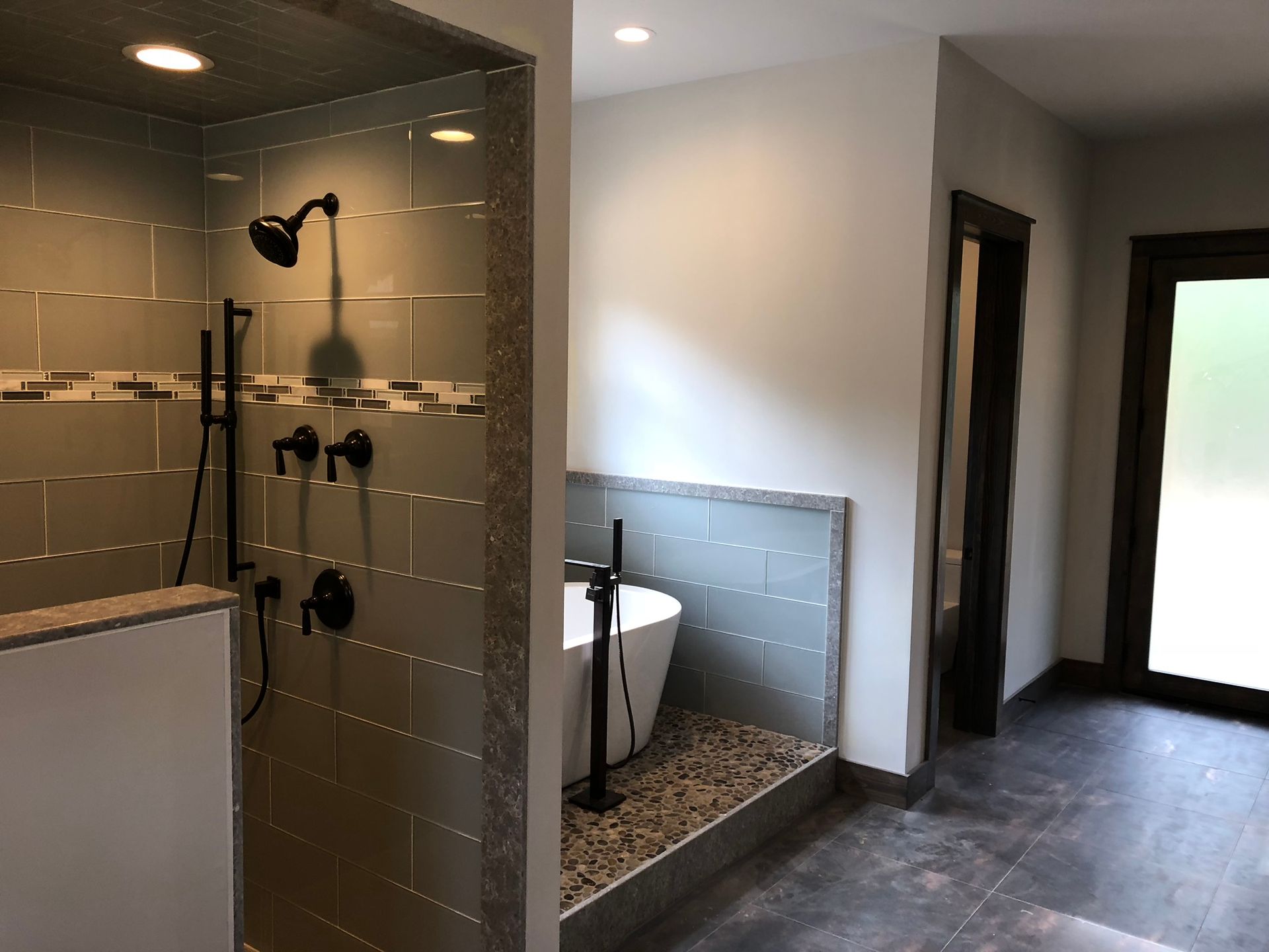 Modern bathroom with glass shower, tiled walls, tub alcove, and hallway doorway beside a bright window.