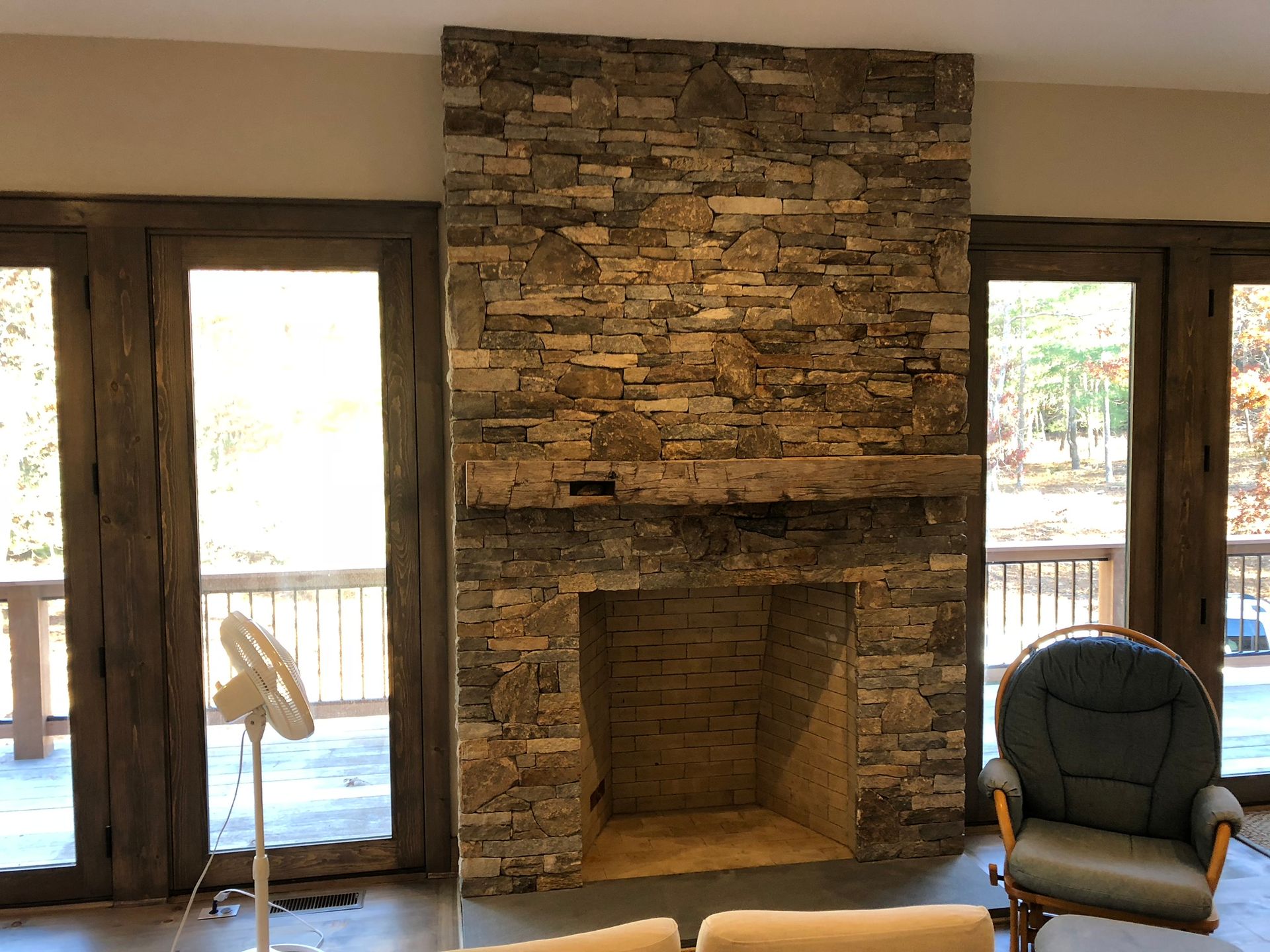 Living room with stone fireplace, large windows, and a blue armchair beside a wooden deck view