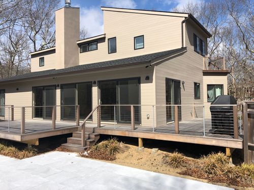 Modern beige house with a raised wooden deck and metal railing, viewed in winter with patches of snow.