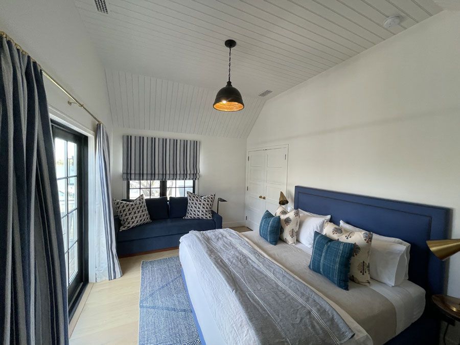 Bright bedroom with a blue bed, striped rug, and couch by large windows and curtains