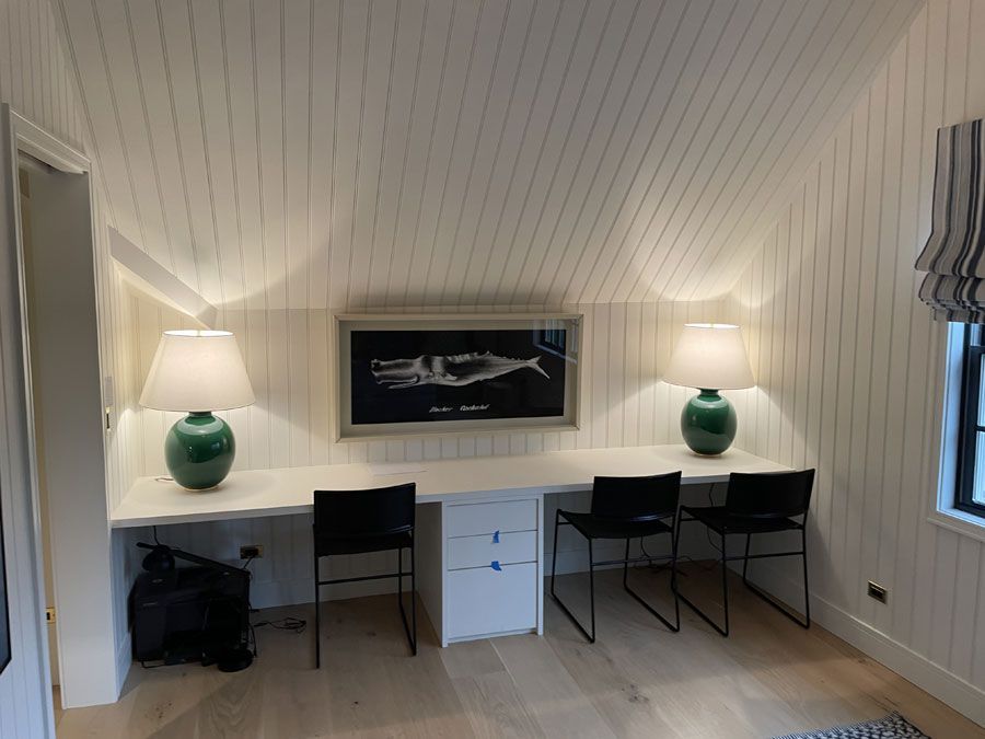 Small home office with two desks, black chairs, green lamps, and a framed picture on a white paneled wall