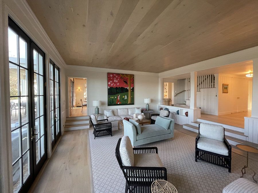 Bright open living room with vaulted wood ceiling, large windows, white sofas, and light wood floors