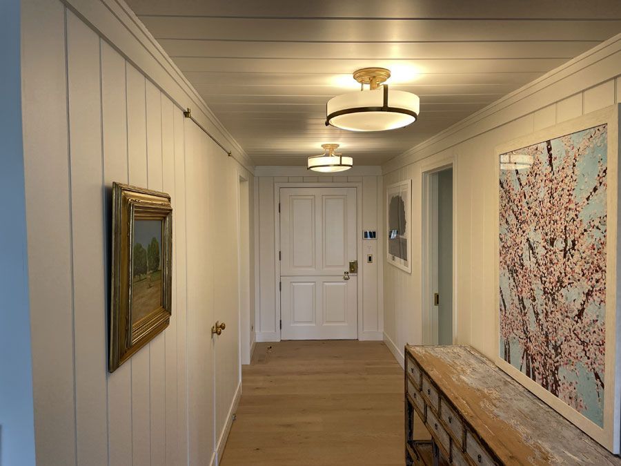 Bright hallway with white paneling, framed art, a wooden console table, and ceiling lights leading to a closed door.