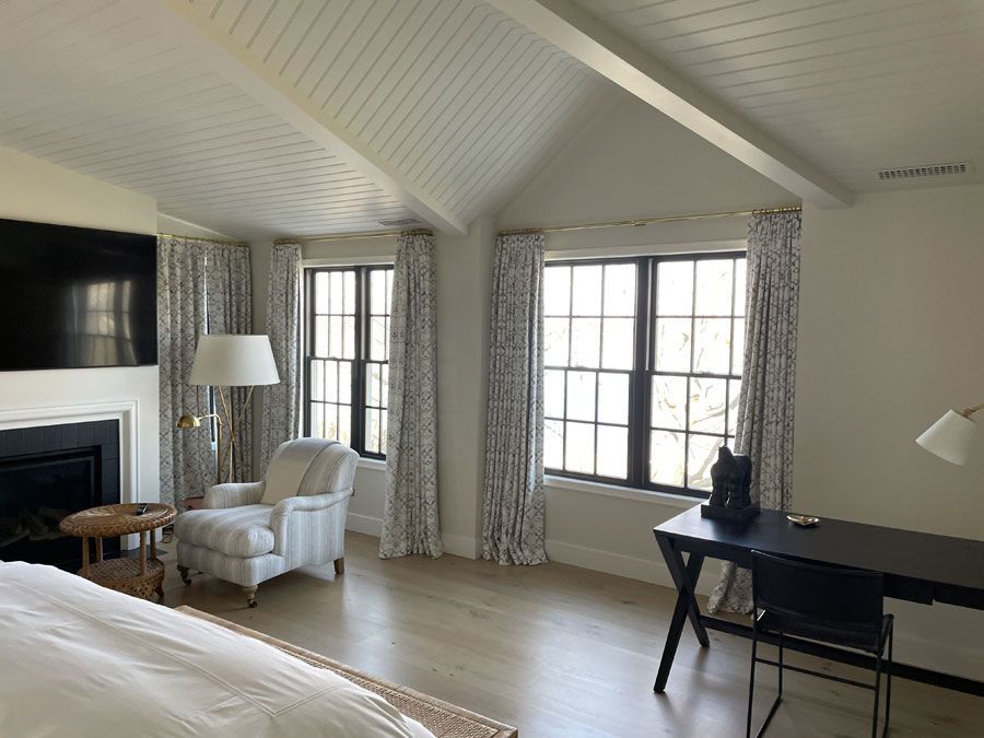 Bright bedroom with vaulted ceiling, large windows, fireplace, armchair, and desk in neutral tones