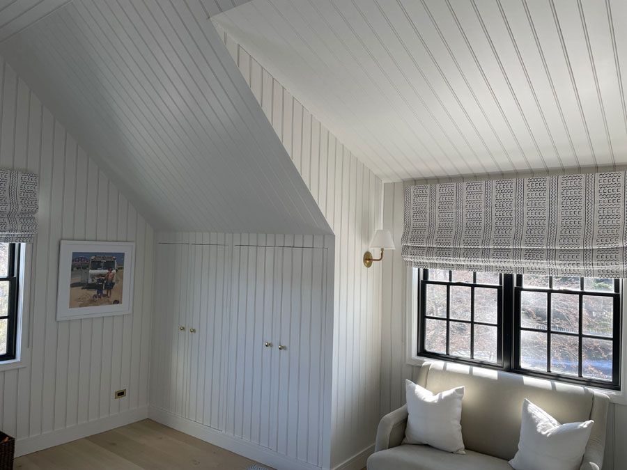 Bright white attic bedroom with sloped ceiling, closet doors, black-framed windows, and a cushioned bench seat