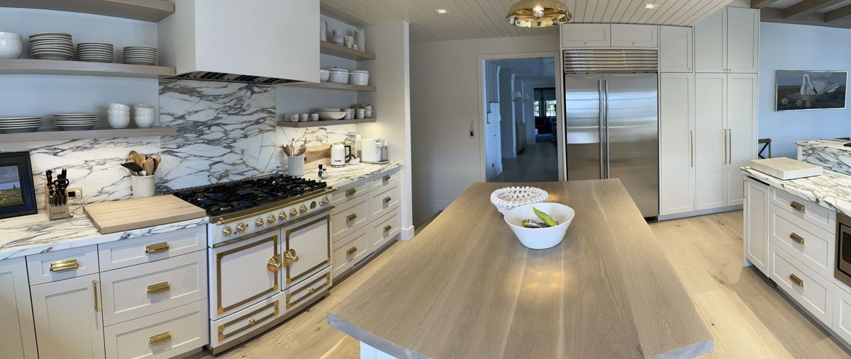 Bright modern kitchen with white cabinets, marble accents, and a long wooden island in the center.