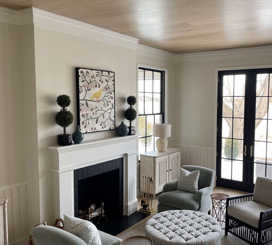 Bright living room with white fireplace, tufted ottoman, armchairs, and French doors.