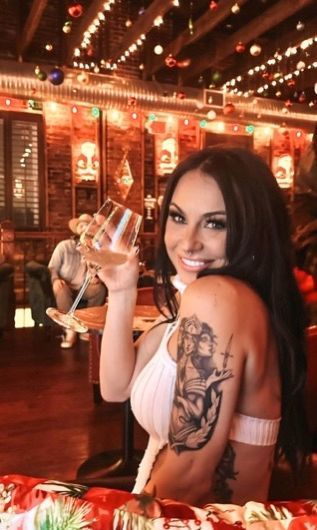 Woman with a tattoo raises a glass of wine, smiling in a festively decorated bar.