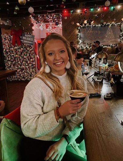 Woman with blonde hair smiles, holding a cocktail in a bar with holiday decorations.