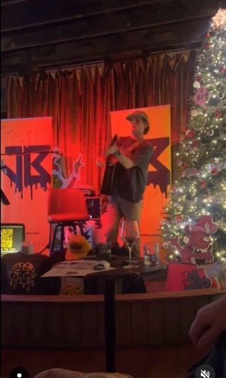 Man on stage with microphone, two orange-lit backdrops, a decorated Christmas tree, and small table.