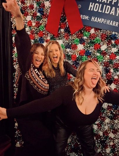 Three women posing playfully in front of a festive wall decorated with bows and tinsel, all smiling with arms raised.