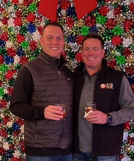 Two men smiling, holding drinks, standing in front of a colorful, festive background.
