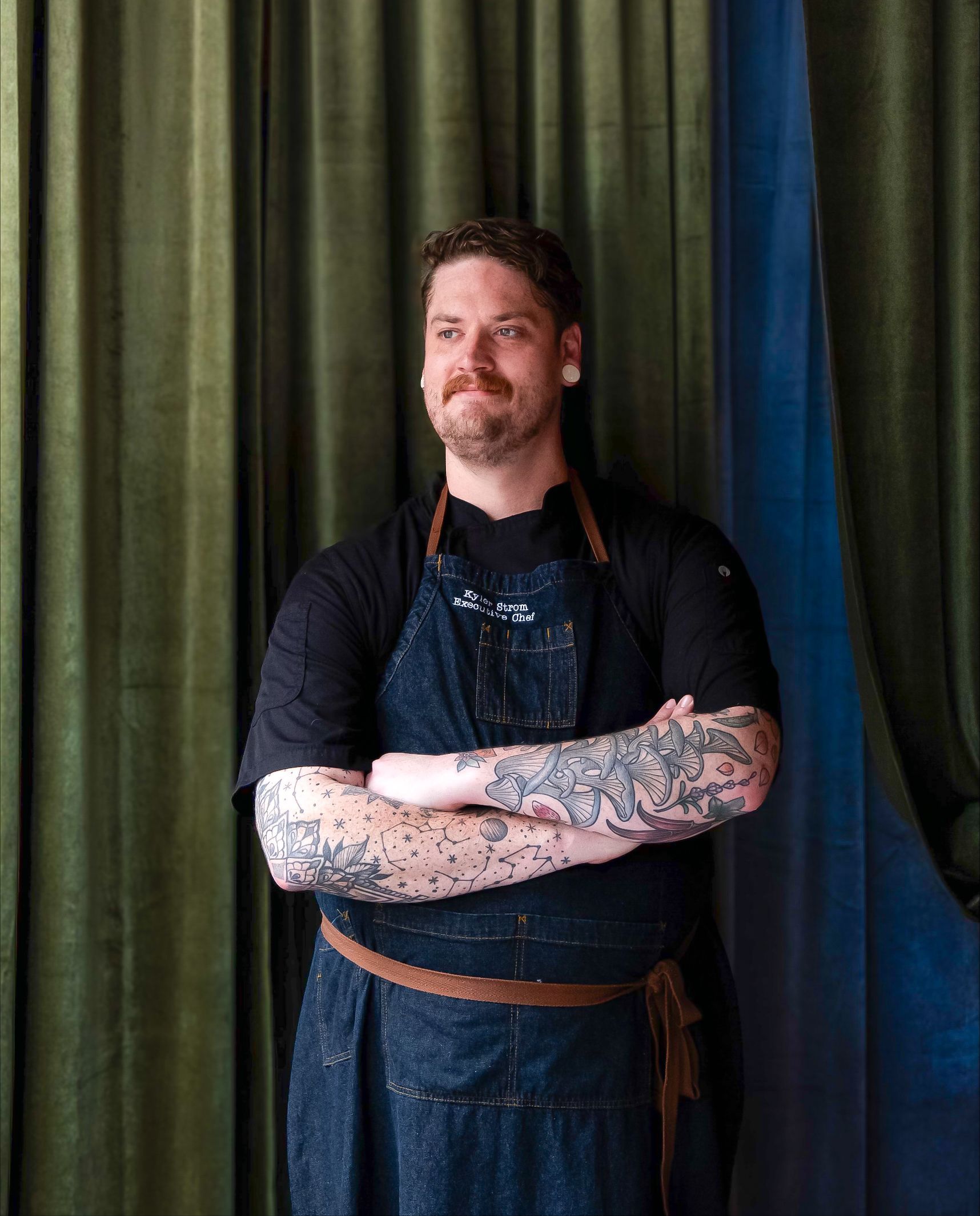 Chef with crossed arms, wearing apron, tattoos, standing in front of green curtains.