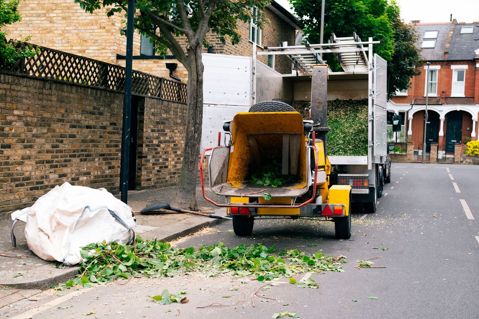 Tree chipper on a street; green branches being processed. White truck and large trash bag next to it.