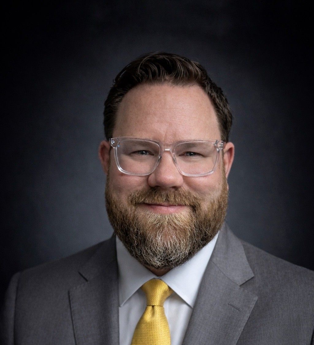 Portrait of a smiling man in a gray suit, yellow tie, and clear glasses on a dark background