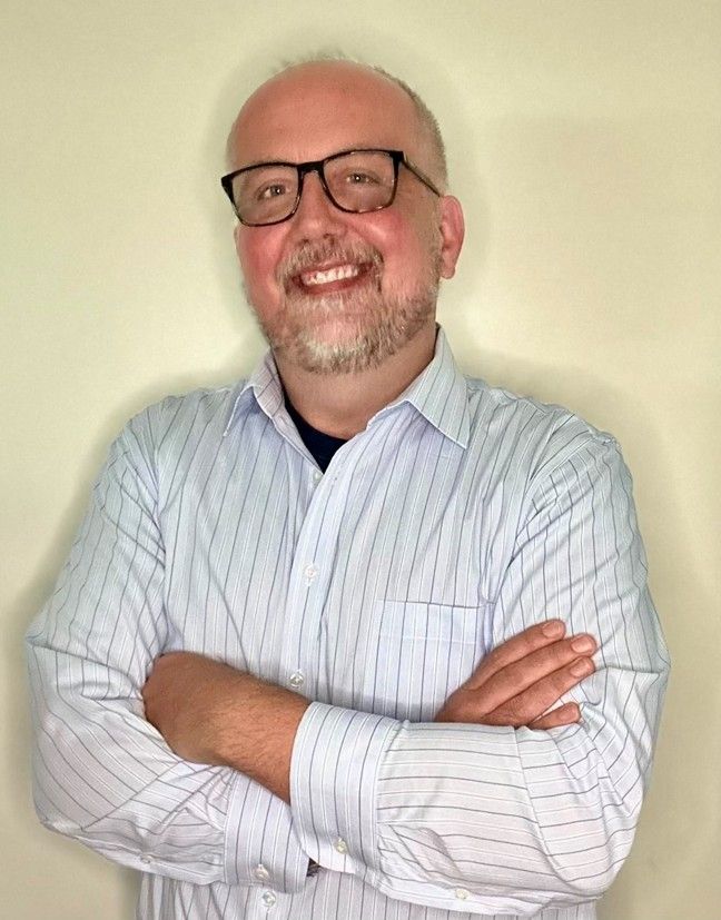 Smiling person in glasses and striped shirt, standing with arms crossed against a plain wall.