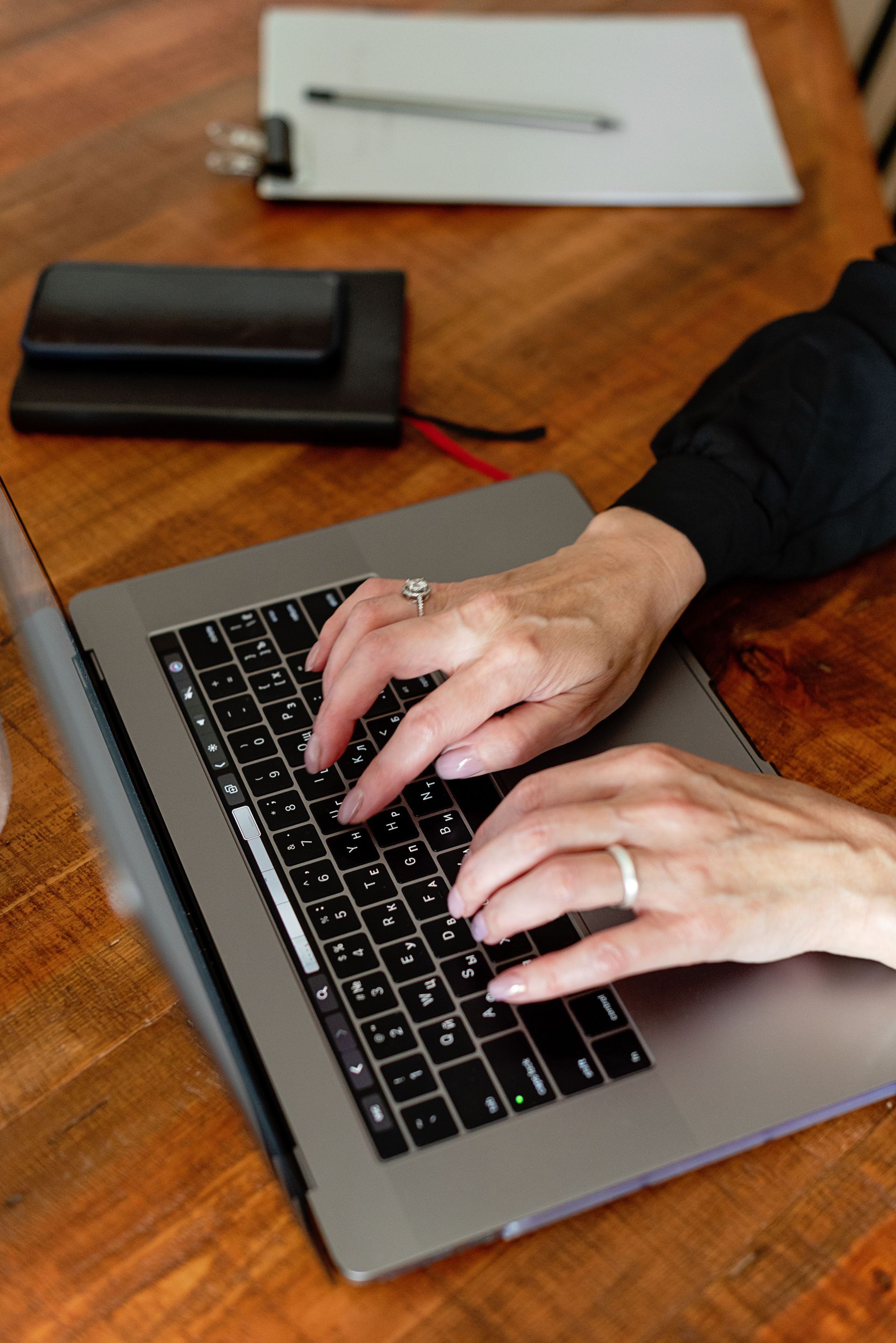 Hands typing on a silver laptop at a wooden desk with a notebook and pen nearby.
