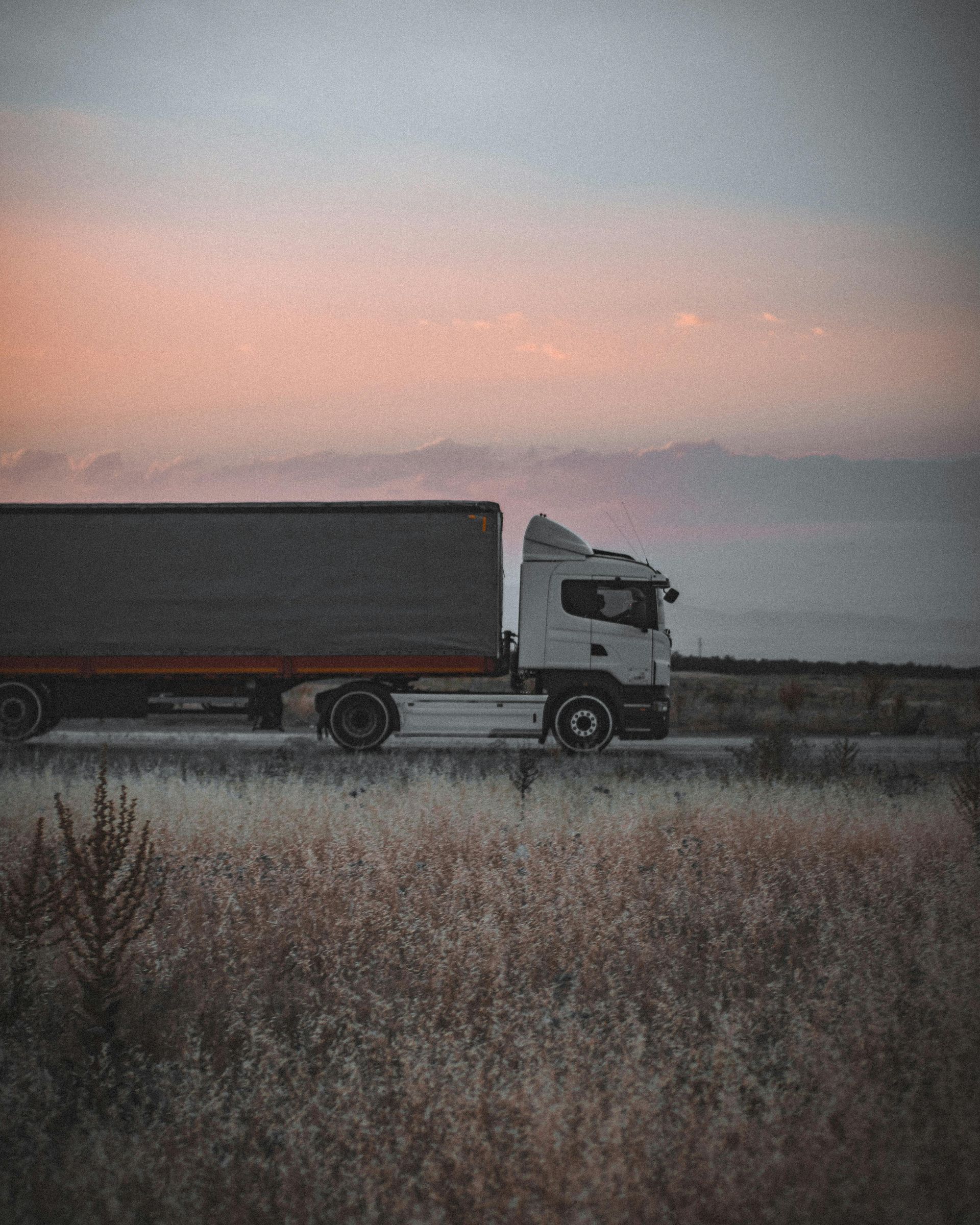 A truck is driving down a road in a field at sunset.