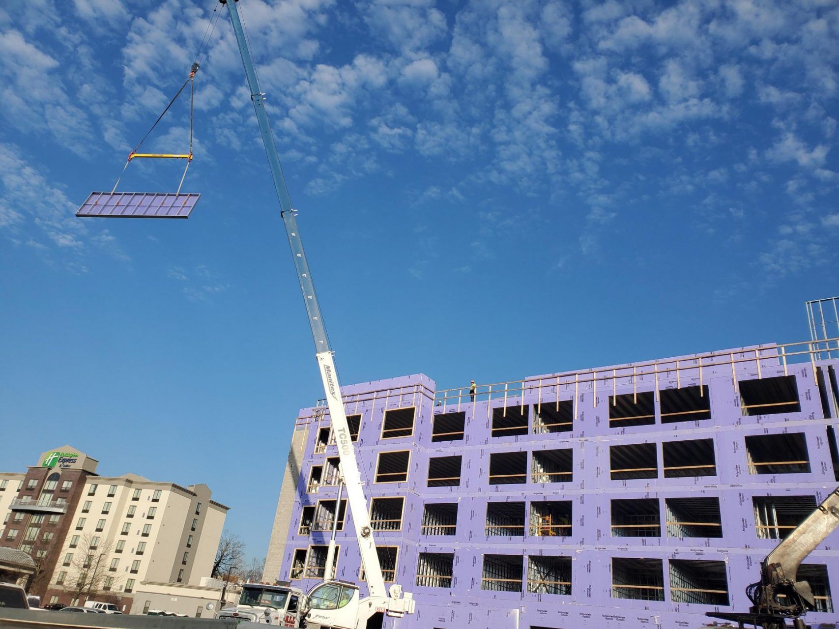 A crane is lifting a large steel section of the building high in the air in front of a large building under construction.