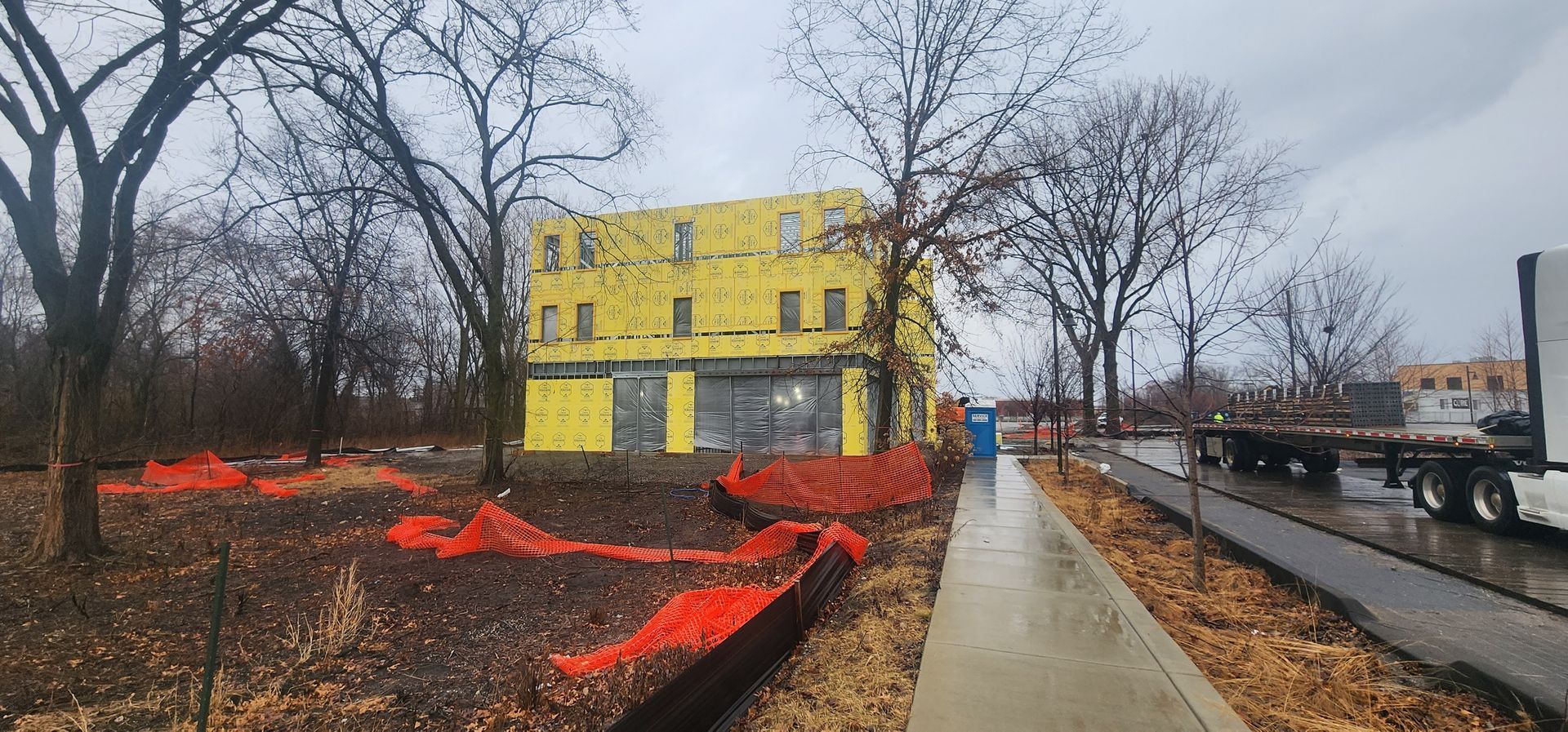 Yellow building beside a wet walkway, with bare trees and orange flower beds under an overcast sky.