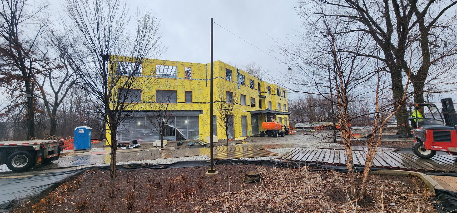 Yellow apartment building under construction with workers, excavator, and bare trees on a rainy day.