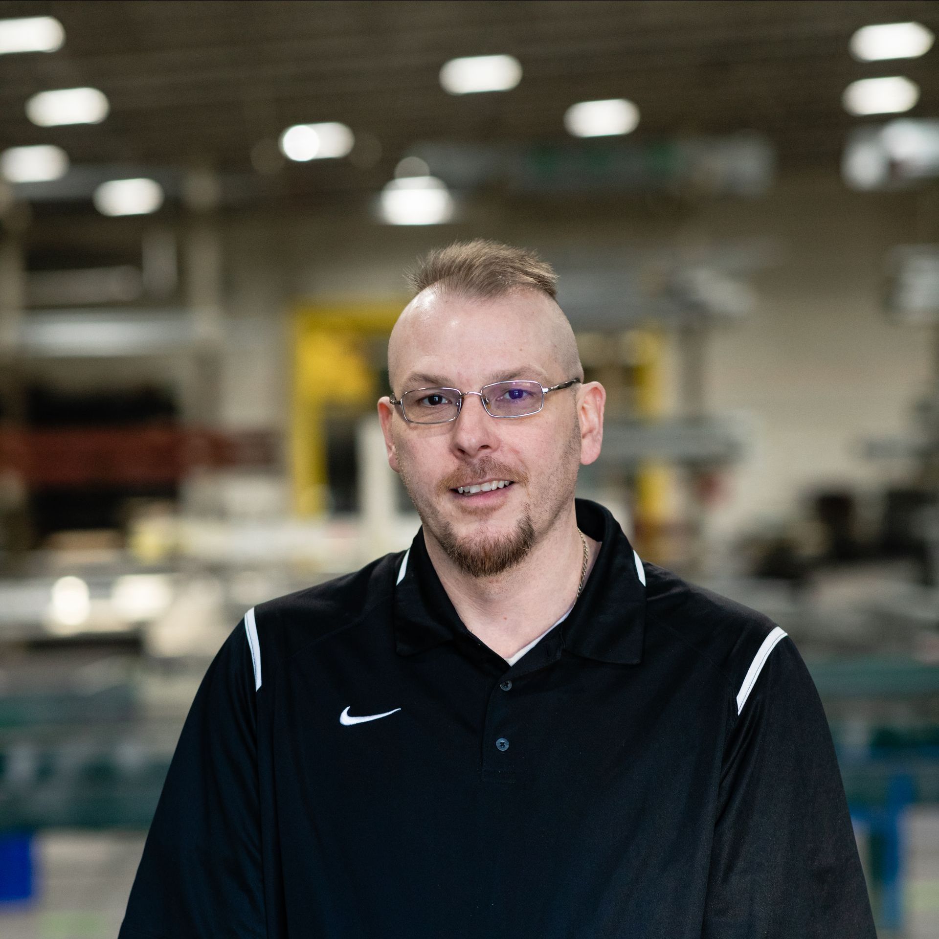 A man wearing glasses and a black shirt is standing in a warehouse and smiling. 