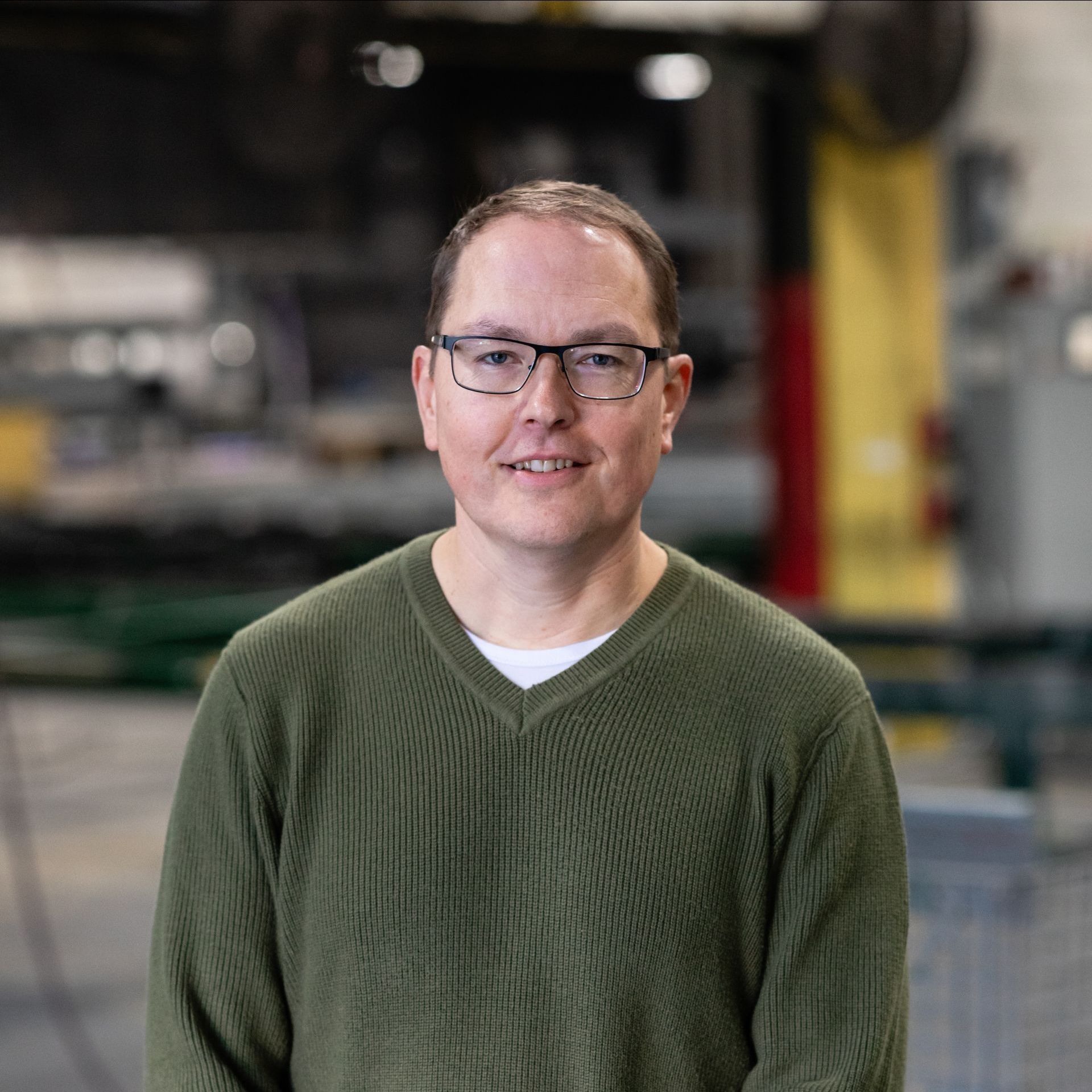 A man wearing glasses and a green sweater is standing in a factory.