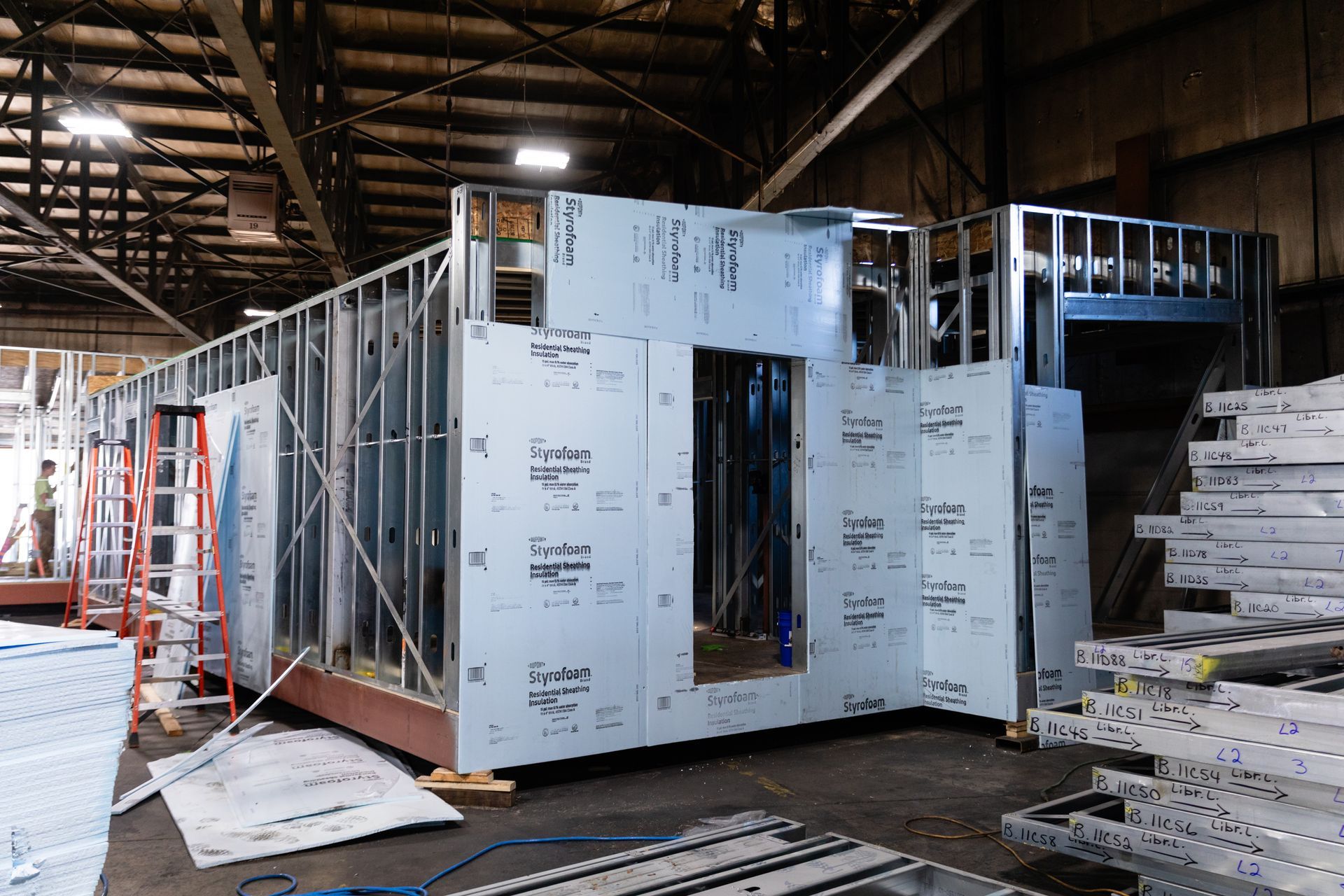 Partially built modular house interior with metal framing and white sheathing in a warehouse.