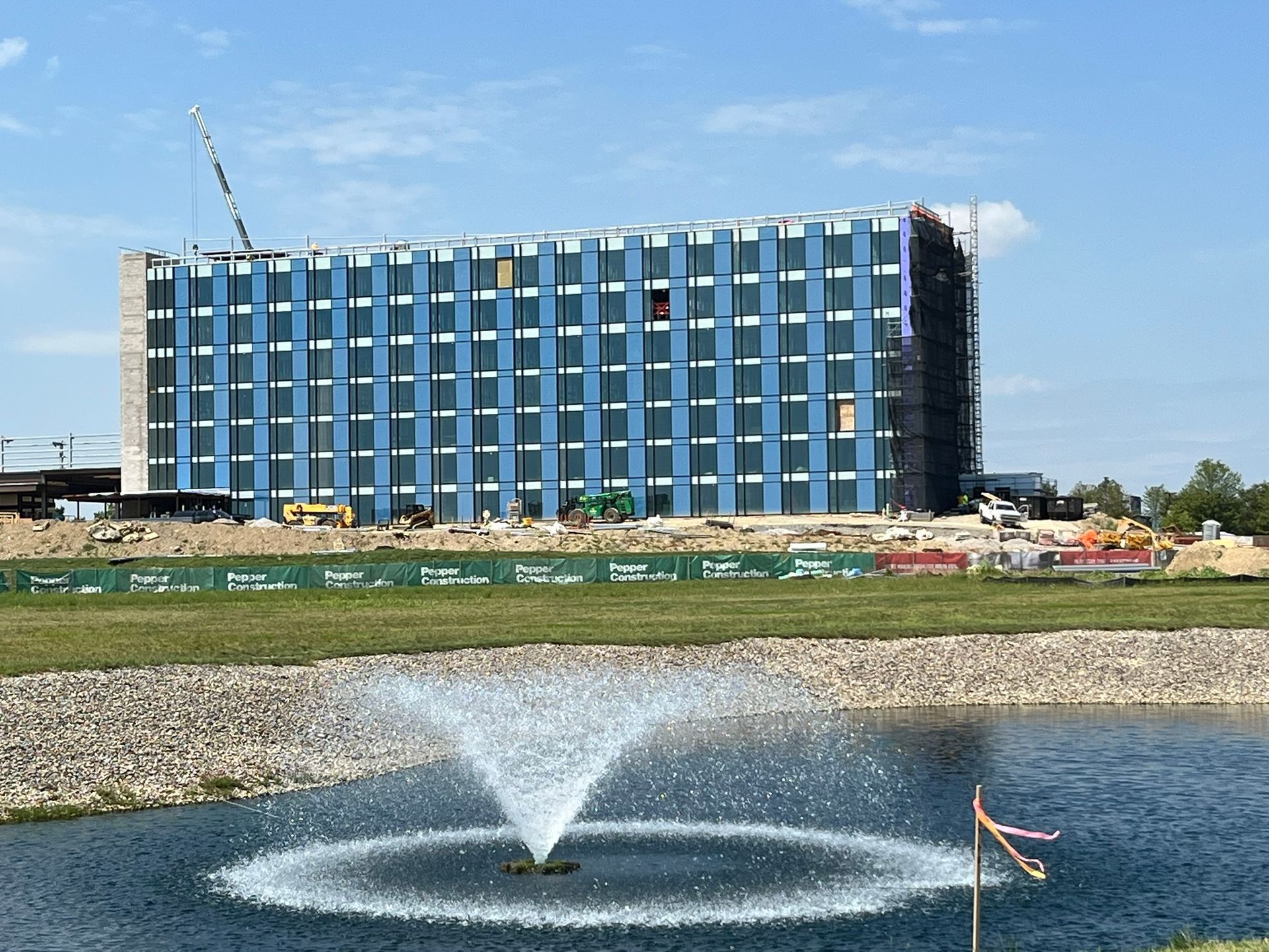 Modern glass building by a pond with a fountain and blue sky.